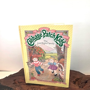 May include: A vintage copy of "Cabbage Patch Kids: The Just-Right Family" storybook. The cover features two children running through a forest scene, with the title in a pink and green banner. The book is a Parker Brothers Story Book.