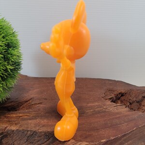 May include: A bright orange plastic figurine of Mickey Mouse. The figurine is standing with its arms at its sides and its head tilted to the side. The figurine is about 6 inches tall.