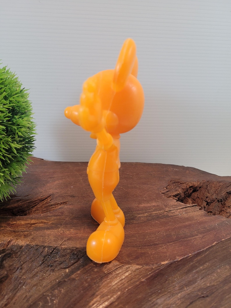 May include: A bright orange plastic figurine of Mickey Mouse. The figurine is standing with its arms at its sides and its head tilted to the side. The figurine is about 6 inches tall.