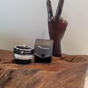 May include: A silver and black Auto 2X Converter lens with a black leather case, and a dark brown wooden peace sign sculpture. The items are displayed on a wooden surface.