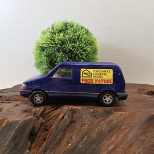 May include: A miniature blue van with the text "Publishers Clearing House Prize Patrol" on the side. The van is parked on a wooden surface, with a green, spherical plant in the background.