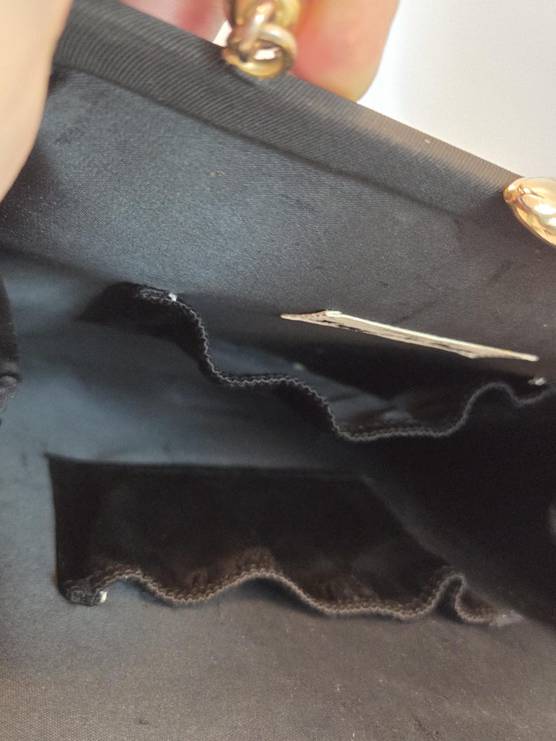 Pode incluir: Bolsa de noite de cetim preto com fecho dourado. O interior apresenta um bolso de veludo preto e uma pequena etiqueta. A bolsa est&aacute; aberta, revelando o forro e os compartimentos.