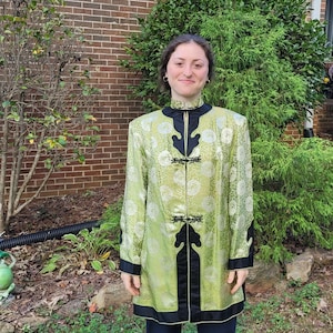 May include: A person wearing a green and black patterned jacket with a mandarin collar and decorative buttons. The jacket has a black trim and a silver lining. The person is standing in front of a brick wall and green bushes.