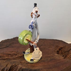 May include: A ceramic clown figurine with a green drum, wearing a gray suit, and a small black top hat. The clown has a red nose and hair, and a tag that reads "T.P. Ceramiche". The figurine is on a yellow base.