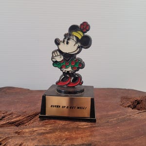 May include: A black and red figurine of Minnie Mouse wearing a green and red dress with white polka dots. The figurine is on a black base with a gold plaque that reads "Hurry Up & Get Well!"