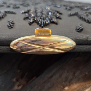 Pode incluir: Close-up de uma bolsa de noite preta com contas e fecho dourado. A bolsa apresenta intrincados bordados florais de contas e uma forma retangular. O fecho &eacute; oval com um design geom&eacute;trico.