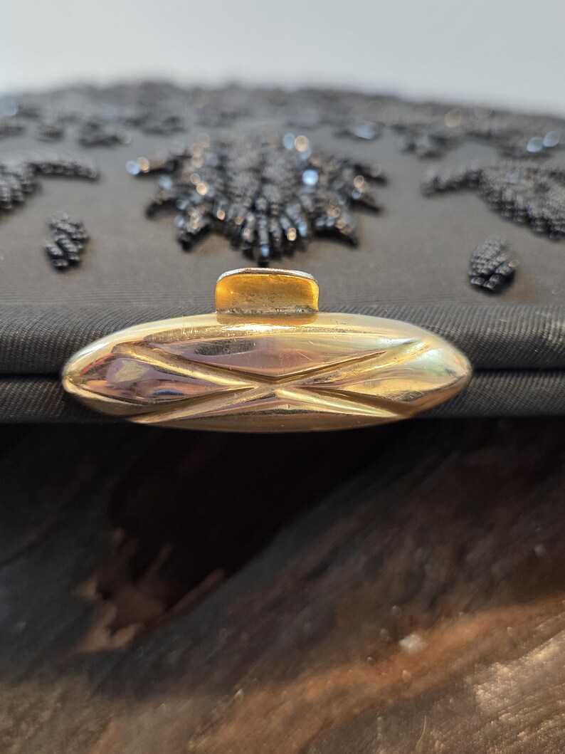 Pode incluir: Close-up de uma bolsa de noite preta com contas e fecho dourado. A bolsa apresenta intrincados bordados florais de contas e uma forma retangular. O fecho &eacute; oval com um design geom&eacute;trico.