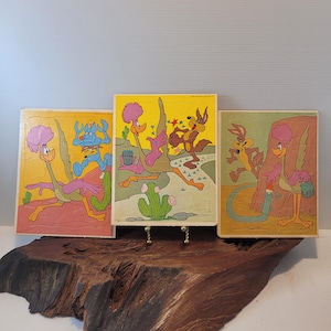 May include: Three rectangular jigsaw puzzles featuring cartoon characters Road Runner and Wile E. Coyote in desert settings. The puzzles display vibrant yellow, pink, and brown backgrounds with various action scenes, including a blue robot character.