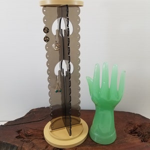 May include: A beige rotating jewelry stand with a black and white patterned design. The stand has three tiers for earrings and a small hand-shaped ring holder.