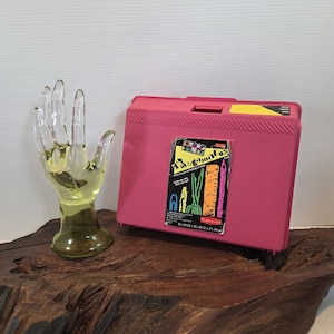May include: A pink plastic storage box with a sticker that reads "TangamaBox" and a clear glass hand sculpture. The box is approximately 9 inches wide, 8 inches deep, and 2 inches high. The hand sculpture is yellow.