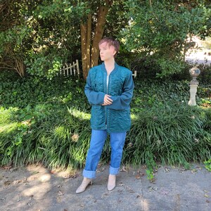 May include: A teal quilted jacket with a zipper closure and a stand-up collar. The jacket is worn by a person with pink hair and blue jeans.