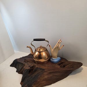 May include: A copper-colored tea kettle with a black handle and brass accents sits on a dark wooden slab. A ceramic hand with red nail polish is displayed on a blue base.