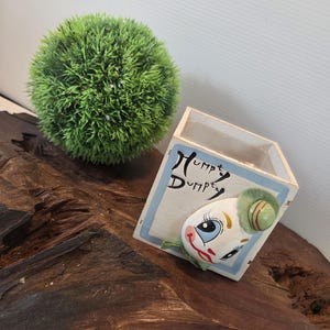 May include: A decorative wooden box with the words "Humpty Dumpty" and a cartoon character face. A green, spherical plant sits next to the box on a wooden surface. The box has a blue border.