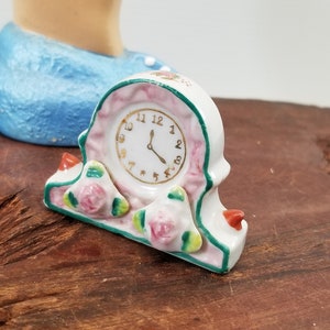 May include: A small white porcelain clock with a pink face and gold numbers. The clock is decorated with green leaves and pink roses. The clock is sitting on a wooden surface.