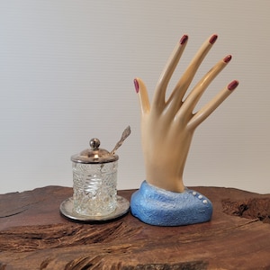 May include: A clear glass jar with a silver lid and a small silver spoon sits on a wooden surface next to a ceramic hand sculpture with red nail polish. The hand is wearing a blue fabric glove with white buttons.