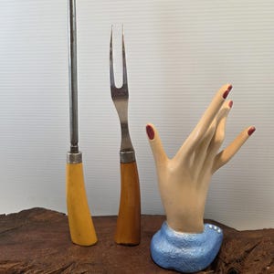 May include: A carving fork and a sharpening steel with wooden handles, next to a ceramic hand sculpture with red nail polish and a blue base. The items are displayed on a wooden surface.