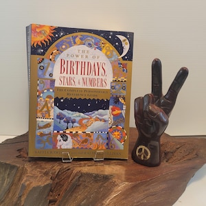 May include: A book titled "The Power of Birthdays, Stars, & Numbers: The Complete Personology Reference Guide" by Saffi Crawford and Geraldine Sullivan, with a colorful cover featuring a sun, moon, and stars, is displayed on a wooden surface next to a brown peace sign hand sculpture.