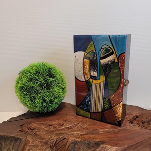 May include: A wooden box with a hinged lid. The lid is decorated with a colorful abstract design featuring geometric shapes and lines in shades of blue, red, yellow, green, and white.