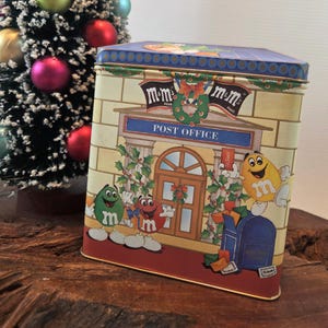 May include: A rectangular M&M's brand tin with a Christmas theme. The tin features a post office scene with M&M's characters, holly, and the words "Post Office". The tin is on a wooden surface with a small Christmas tree in the background.
