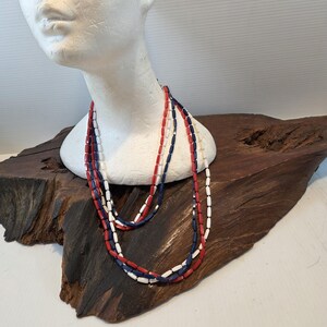 May include: A multi-strand necklace featuring cylindrical beads in alternating red, white, and navy blue colors. The long, layered necklace consists of several strands of uniform, smooth, tubular-shaped beads strung together in a continuous loop.