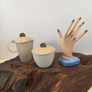 May include: A cream-colored teacup with a handle and a matching lidded sugar bowl sit next to a small, round lidded container. A hand-shaped display stand with red painted nails and a blue base is also present.