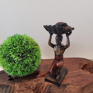 May include: A bronze sculpture of a person kneeling and holding a large seashell above their head. The sculpture is on a wooden base.