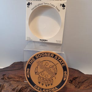 May include: A round Frankoma terra cotta trivet featuring a dark blue border and a central black graphic of an Oklahoma state emblem with a peace pipe and feathers. The text reads THE SOONER STATE OKLAHOMA. It is displayed with its original white box.