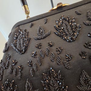 Pode incluir: Bolsa preta com contas com uma al&ccedil;a escura e ferragens douradas. A bolsa apresenta um padr&atilde;o repetido de desenhos de folhas com contas. A al&ccedil;a &eacute; marrom escuro com detalhes dourados.