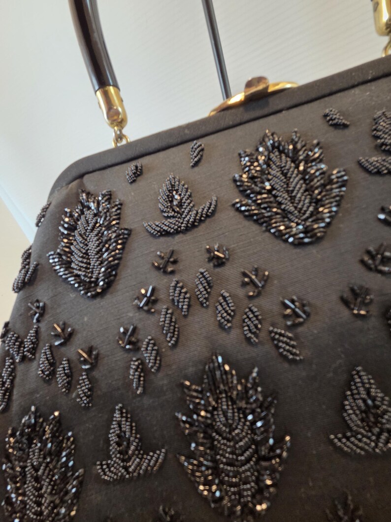 Pode incluir: Bolsa preta com contas com uma al&ccedil;a escura e ferragens douradas. A bolsa apresenta um padr&atilde;o repetido de desenhos de folhas com contas. A al&ccedil;a &eacute; marrom escuro com detalhes dourados.