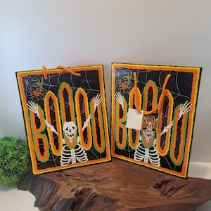 May include: Two rectangular black gift bags featuring a skeleton graphic with arms raised. The word BOOO is printed in large orange letters with green outlines. The background includes white spiderwebs and orange spiders. Each bag has orange rope handles.
