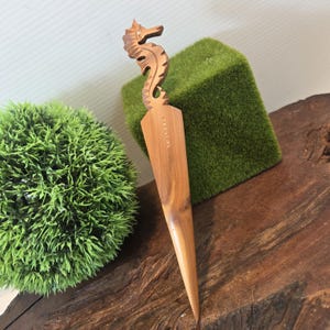 May include: A wooden letter opener with a seahorse carving at the top. The opener is light brown and tapers to a point. The seahorse is intricately carved with details. The word "Sazare" is etched on the opener.