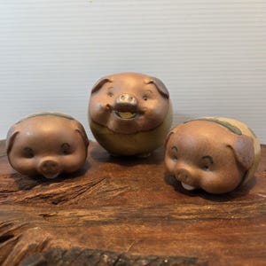 May include: Three ceramic pig figurines with a smooth, matte brown finish on their faces and ears. The central pig is larger and rounder, while the two smaller pigs on either side have a flatter, more compact shape with visible snout details.