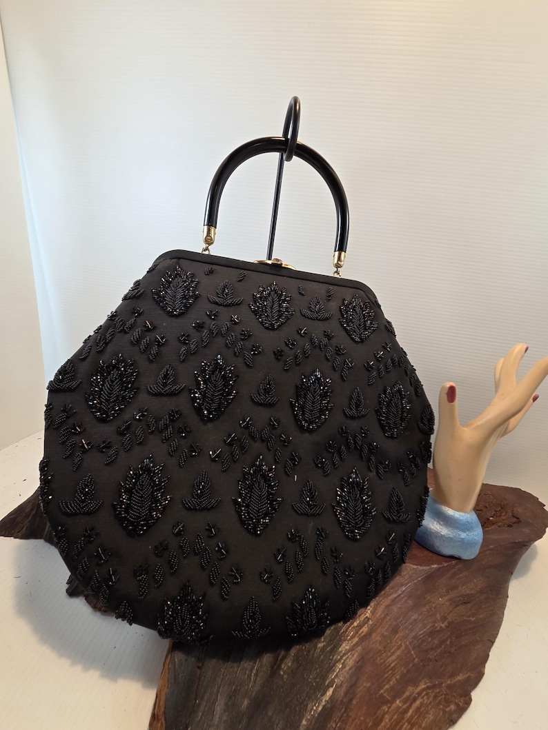 Pode incluir: Uma bolsa preta com contas, com formato arredondado e al&ccedil;a preta. A bolsa &eacute; coberta por intrincados trabalhos de contas, com padr&otilde;es florais e de folhas. A al&ccedil;a tem um design curvo.
