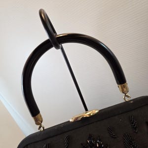 Pode incluir: Uma mala preta vintage com uma al&ccedil;a curva e brilhante. A mala apresenta um design de folhas com contas e ferragens douradas. A al&ccedil;a &eacute; presa &agrave; mala com metal dourado. A mala &eacute; exibida em um suporte de metal preto.