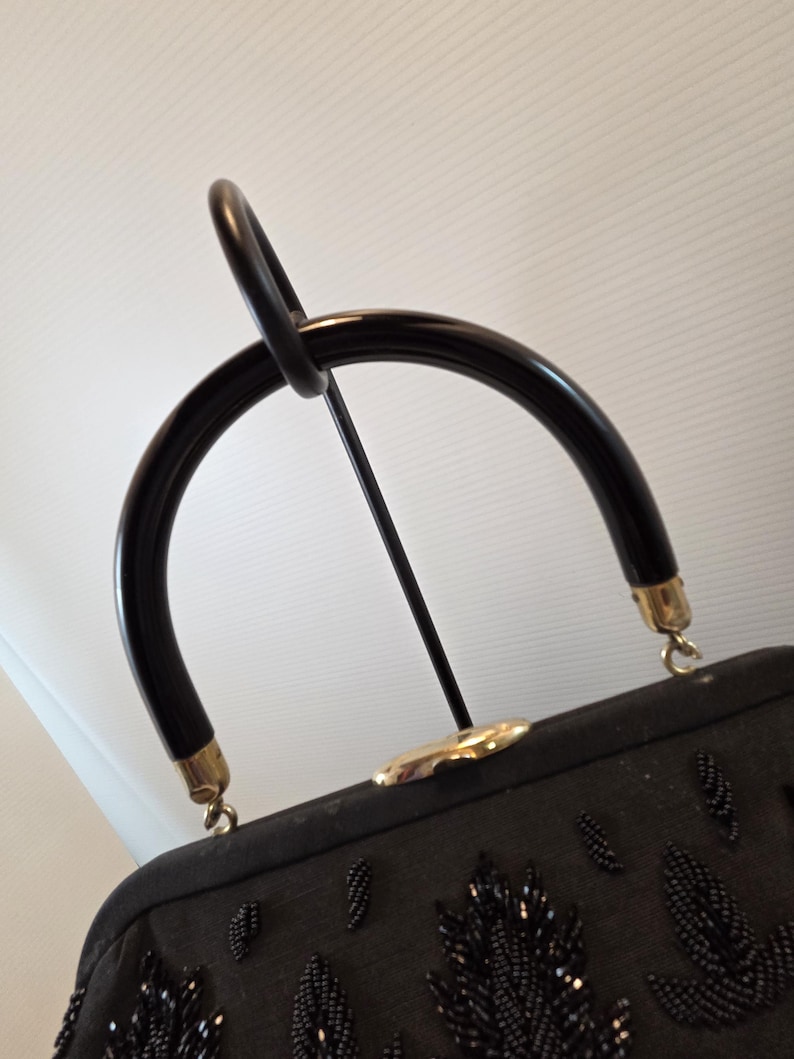 Pode incluir: Uma mala preta vintage com uma al&ccedil;a curva e brilhante. A mala apresenta um design de folhas com contas e ferragens douradas. A al&ccedil;a &eacute; presa &agrave; mala com metal dourado. A mala &eacute; exibida em um suporte de metal preto.