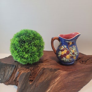 May include: A blue and brown ceramic pitcher with a floral design sits on a wooden surface next to a green ball of artificial grass.