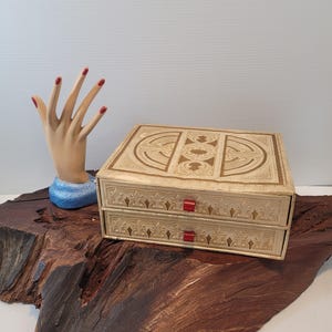 May include: A vintage two-drawer jewelry box with a cream-colored finish and ornate embossed designs. The box features red rectangular drawer pulls. A mannequin hand with red nail polish is on the left side of the box.