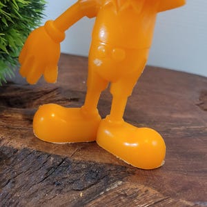 May include: Orange plastic toy figurine of a cartoon character with a large head, short legs, and large hands. The figurine is standing on a wooden surface.