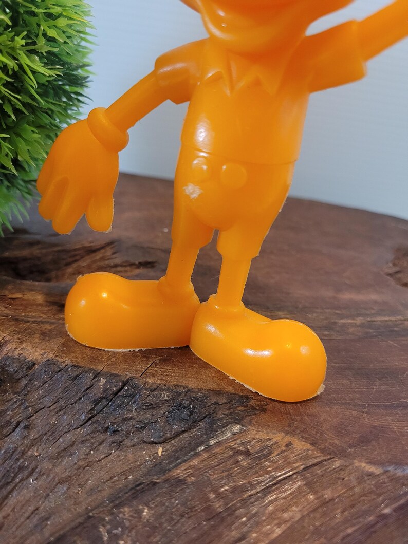 May include: Orange plastic toy figurine of a cartoon character with a large head, short legs, and large hands. The figurine is standing on a wooden surface.