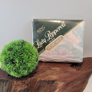 May include: A packaged King flat sheet by Lady Pepperell. The package is dark green with white text and a floral pattern. The sheet is labeled as no-iron percale and made in the USA. A green decorative ball is to the left.