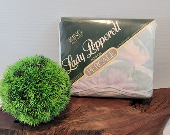 May include: A packaged King flat sheet by Lady Pepperell. The package is dark green with white text and a floral pattern. The sheet is labeled as no-iron percale and made in the USA. A green decorative ball is to the left.