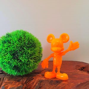 May include: Orange plastic Mickey Mouse figurine with a raised hand, standing on a wooden surface next to a green ball of artificial grass.