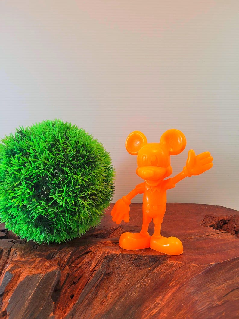 May include: Orange plastic Mickey Mouse figurine with a raised hand, standing on a wooden surface next to a green ball of artificial grass.