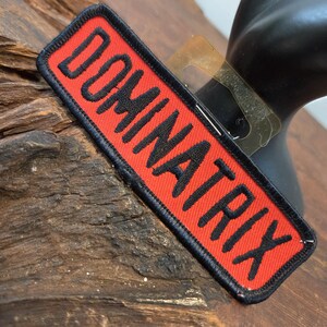 May include: A rectangular embroidered patch featuring a bright red background with a thick black border. The word DOMINATRIX is stitched in bold black capital letters across the center of the patch, oriented diagonally from the bottom left to top right.