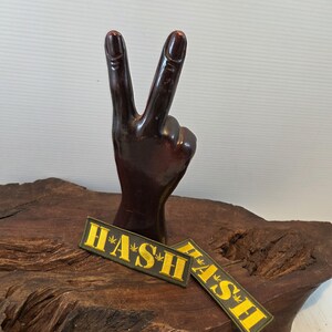 May include: A dark brown wooden hand sculpture shaped in a peace sign gesture. Two rectangular fabric patches are positioned in front, featuring a dark olive green background with the yellow text H*A*S*H and small star-like symbols between letters.