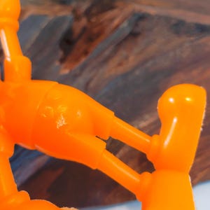 May include: Orange plastic toy figure of a cartoon character with large feet and hands. The figure is posed with its arms outstretched and legs bent.