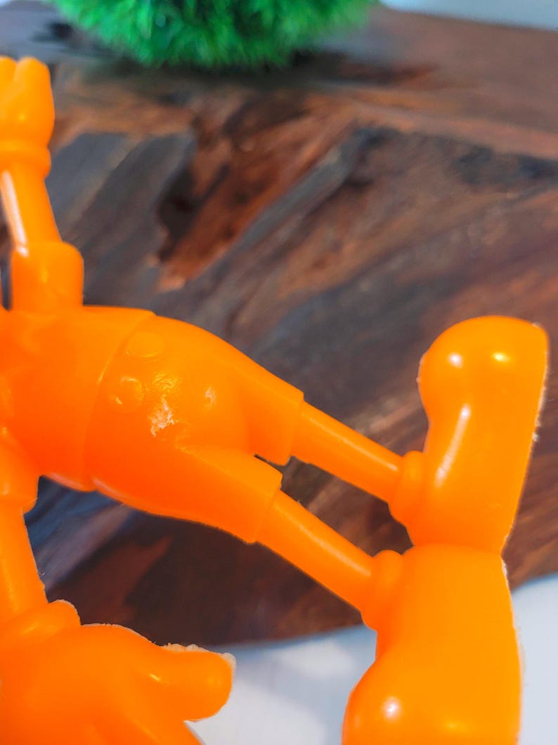 May include: Orange plastic toy figure of a cartoon character with large feet and hands. The figure is posed with its arms outstretched and legs bent.