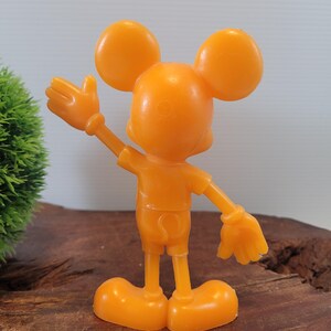 May include: Orange plastic Mickey Mouse figurine with a raised hand, standing on a wooden surface.