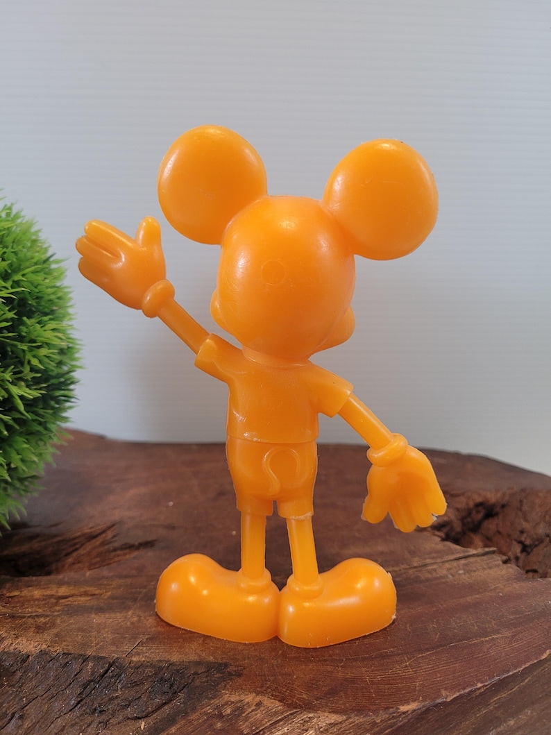 May include: Orange plastic Mickey Mouse figurine with a raised hand, standing on a wooden surface.