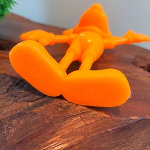 May include: Orange plastic figurine of a cartoon character with large feet and outstretched arms.